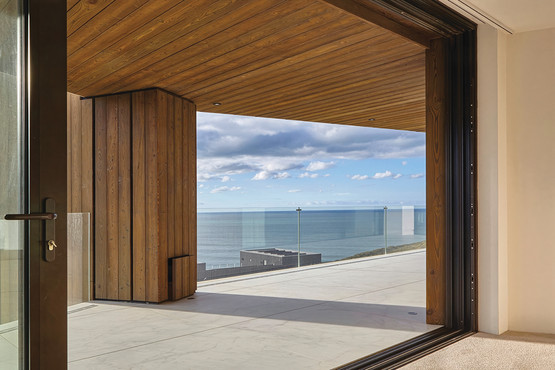 Modern patio with wooden ceiling, glass railing, and ocean view under a blue sky with clouds, seen through an open glass door.