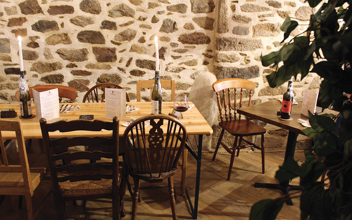 Cozy stone-walled restaurant with wooden tables set for dining. Candles, wine, and menus create a warm, inviting atmosphere.