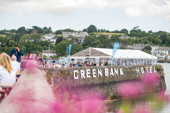 People gather near a tent by a riverside at Greenbank Hotel. Pink flowers in the foreground and houses on a green hill in the background.