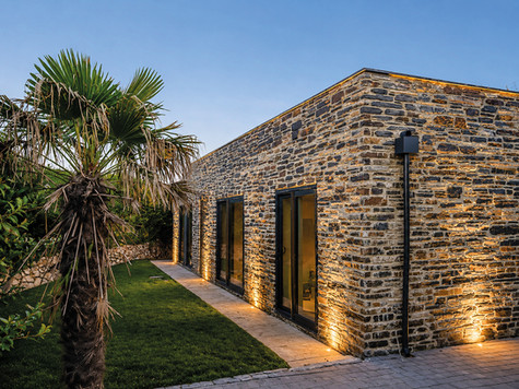 Modern stone house exterior at dusk, lit by warm ground lights beside a palm tree and green lawn.