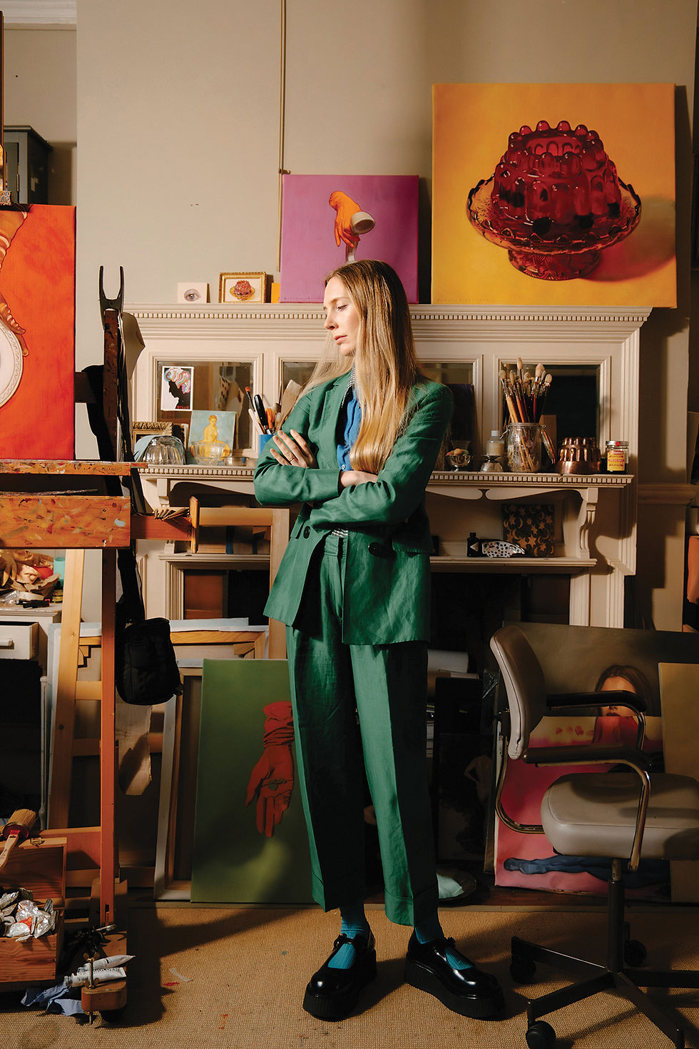 Florence Houston studio 2026 Woman in green suit stands in a cluttered art studio, arms crossed, surrounded by colorful paintings and brushes.
