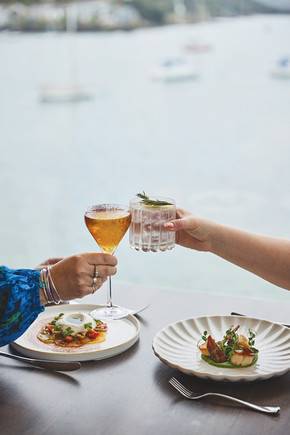 Two hands clink cocktail glasses over a table with elegant plated dishes. Bright window view with boats in the background suggests a coastal setting.