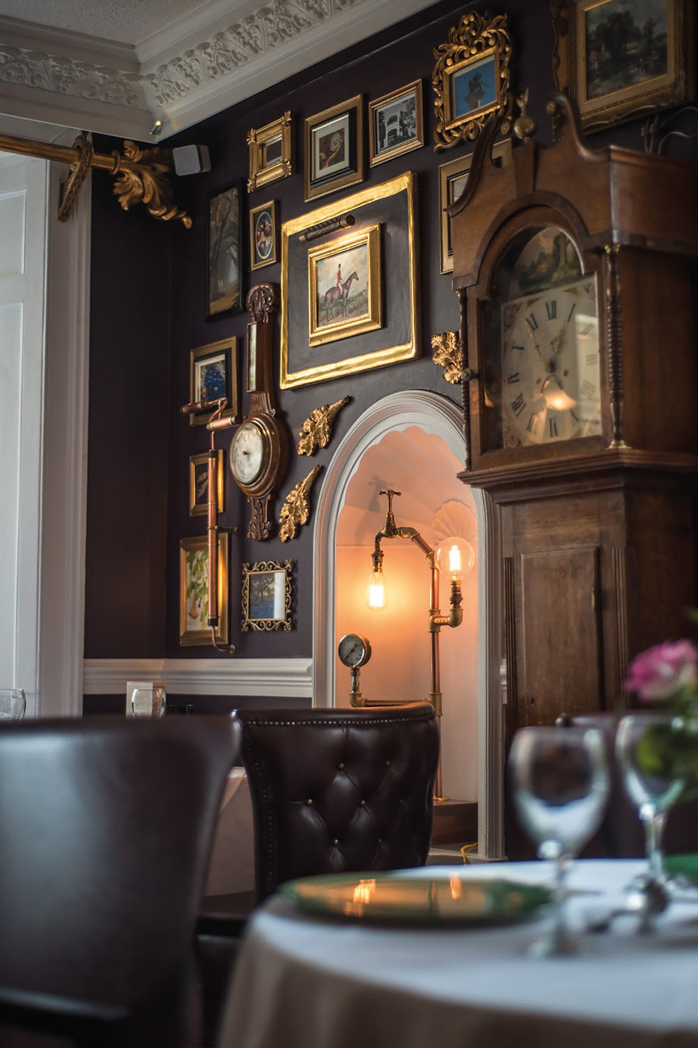 Elegant room with dark walls, vintage clock, framed art, and brass lamp. Leather chairs and set table in foreground, creating a cozy feel.