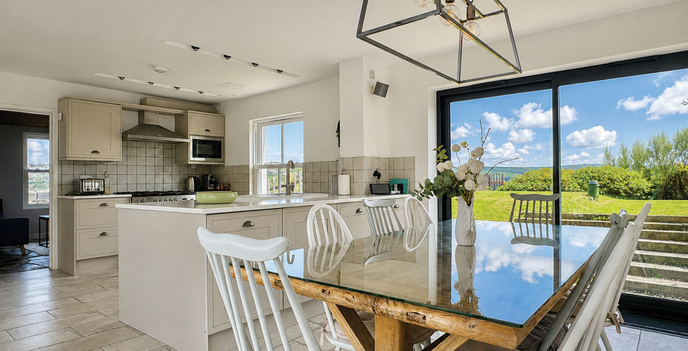 Bright modern kitchen-dining room with glass table, white chairs, and large window showing blue sky and green garden.