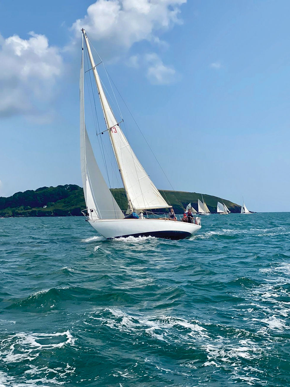 White sailboat with crew and number 3 sails on choppy blue sea near a green island, with other sailboats behind.