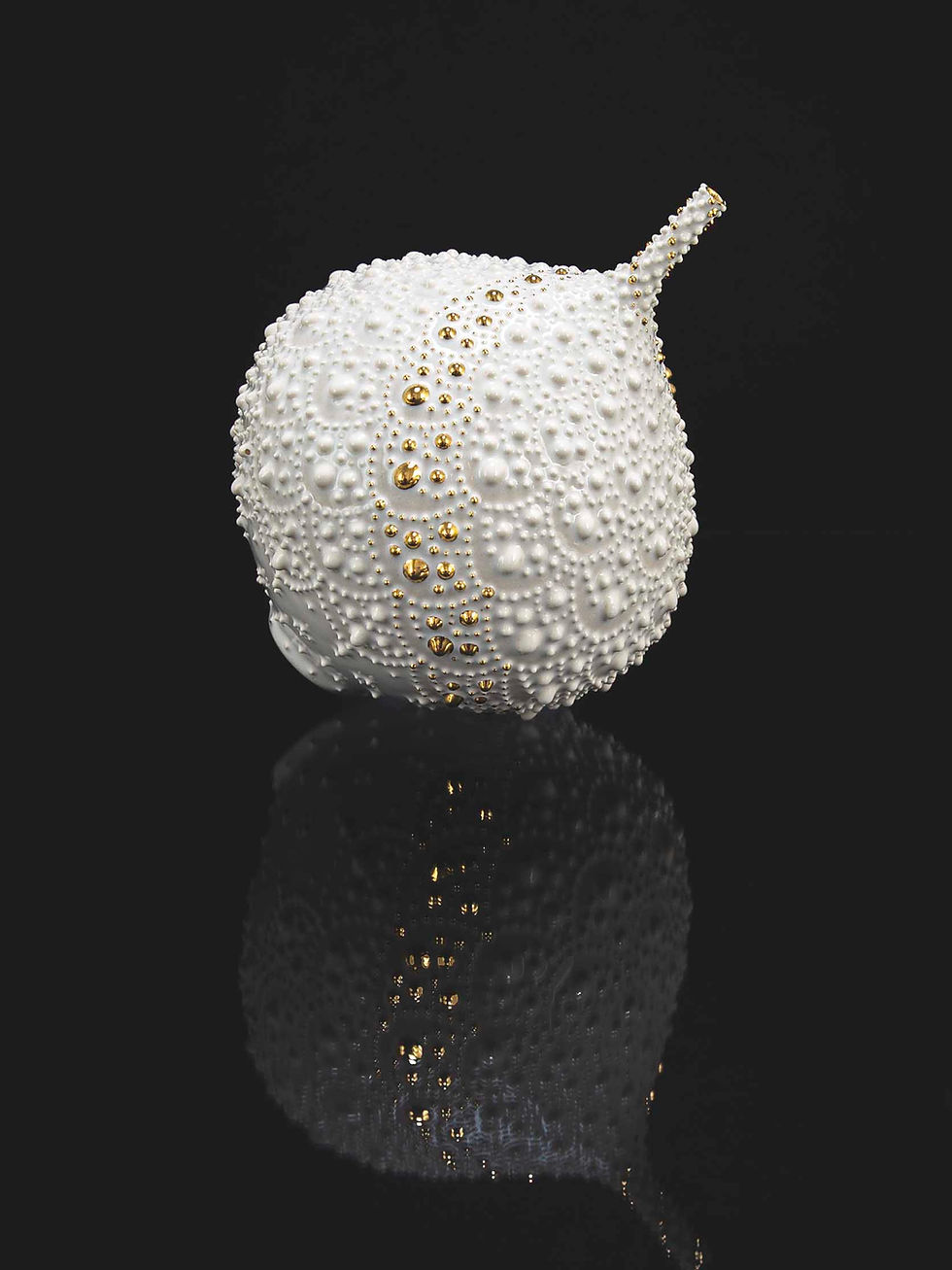 White textured apple-like sculpture with gold beads on a black reflective surface, minimalist studio shot