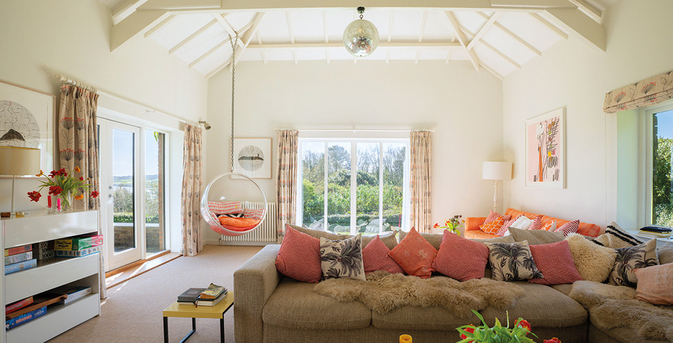 Bright living room with vaulted ceiling, floral curtains, beige sofa with colorful pillows, hanging chair, books, and vibrant tulips on a table.