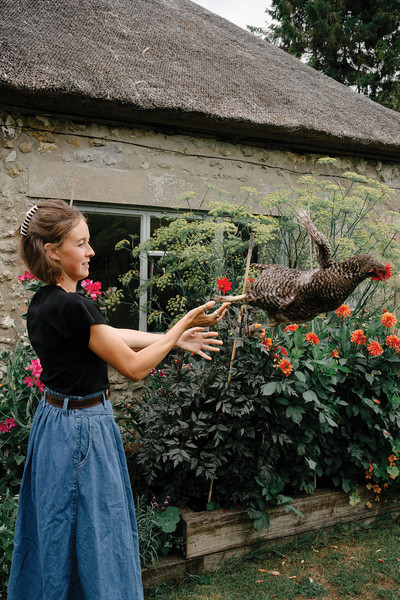 Woman in a blue skirt reaches toward a flying hen in a flower-filled cottage garden.