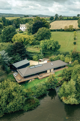 Aerial view of a modern house beside a river in lush countryside with fields, trees, and cloudy skies.