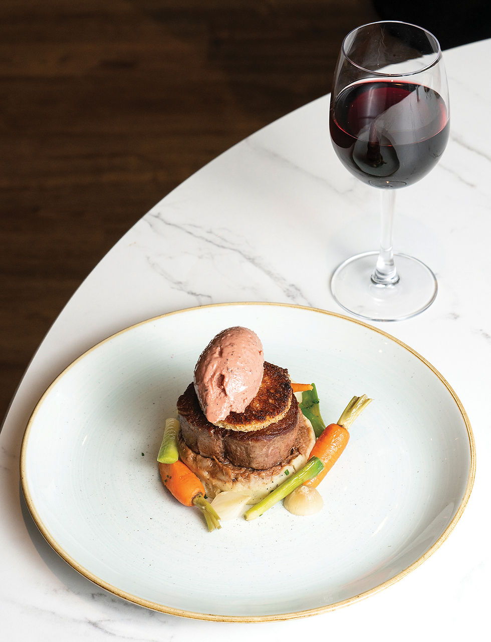 Steak topped with mousse, paired with vegetables on a white plate, next to a glass of red wine on a marble table. Elegant dining scene.