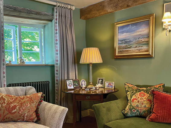 Cozy green living room with open window, lit lamp, framed beach painting, patterned sofas, and family photos on a side table.
