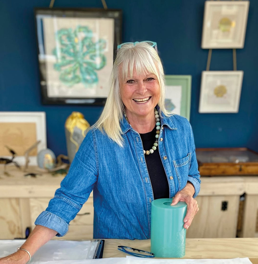 Smiling blonde woman in denim shirt holds a turquoise roll in an art studio with framed artwork on blue walls.
