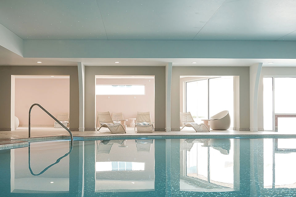 the Indoor pool at the St Moritz Hotel in cornwall with calm water reflects a modern, minimalistic space with beige lounge chairs and large windows, creating a serene ambiance. 