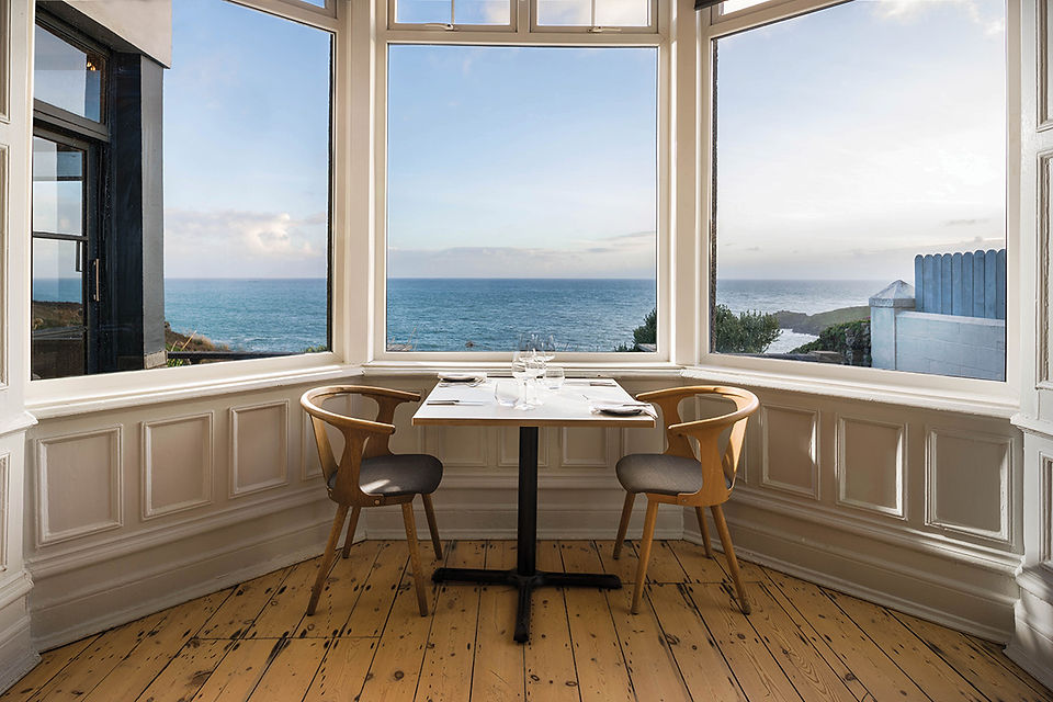 A table set for two by large windows overlooks a scenic ocean view. The room has natural wood flooring and white paneled walls.