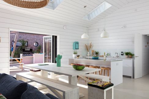 Bright white open-plan kitchen and dining area with wooden table, pendant lights, cactus decor, and patio grill outside.