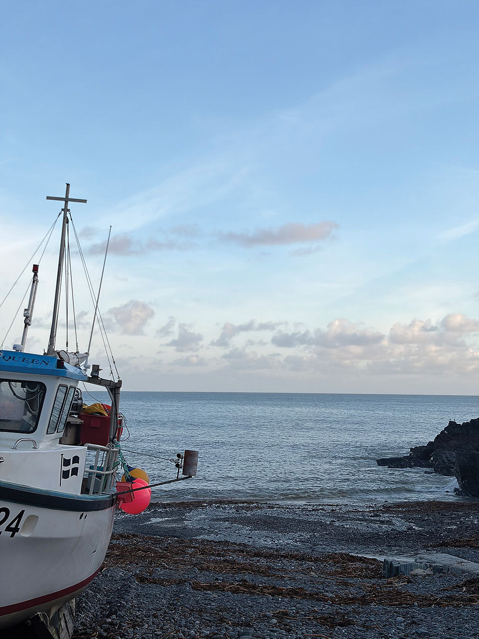 Boat on rocky shore with vibrant buoys, against a calm sea and clear blue sky. Cliff to the right. Text visible on boat.