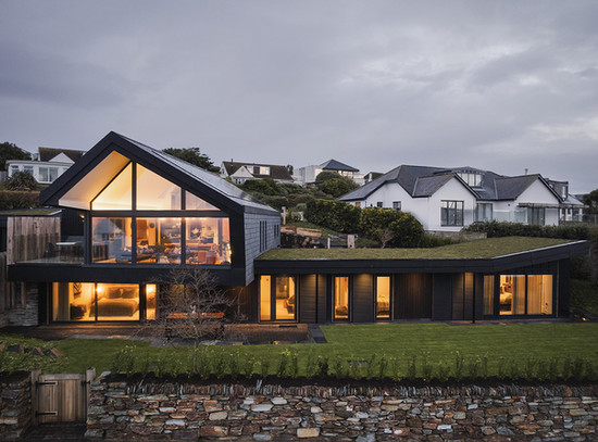 Modern house with large glass windows glowing warmly, surrounded by grass and stone wall. Overcast sky with neighboring houses in view.