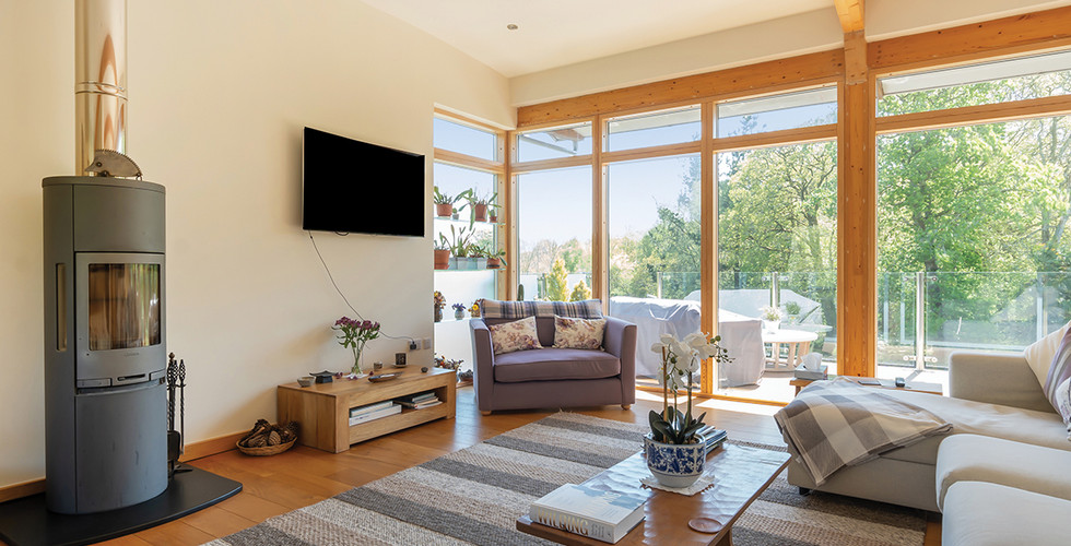 Modern living room with a cozy sofa, wood stove, and TV. Large windows reveal a sunny garden. Neutral tones and floral accents create a calm vibe.