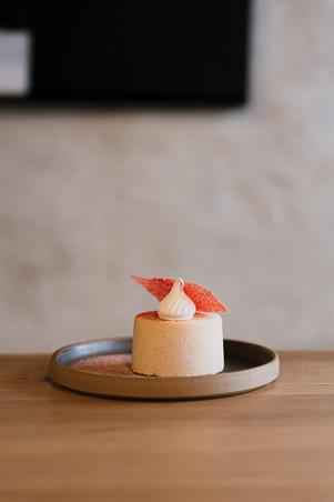 A small, elegant dessert with white icing and a red wafer sits on a beige plate, against a blurred neutral background on a wooden table.