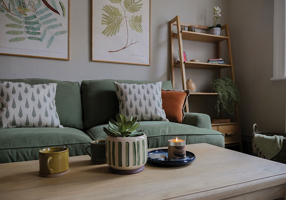 Cosy living room with a green sofa, patterned cushions, botanical wall art, a wooden coffee table and a lit candle.