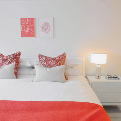 White bed with coral-patterned pillows and throw in a bright bedroom. "Sand Dunes" art on the wall. Lamp and books on bedside table.