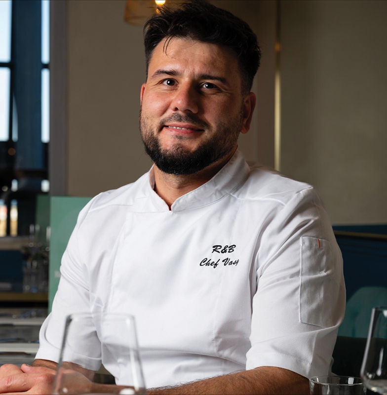 Chef in a white uniform labeled "R&B Chef Vasi" smiles in a restaurant setting with soft lighting and a blurred background.