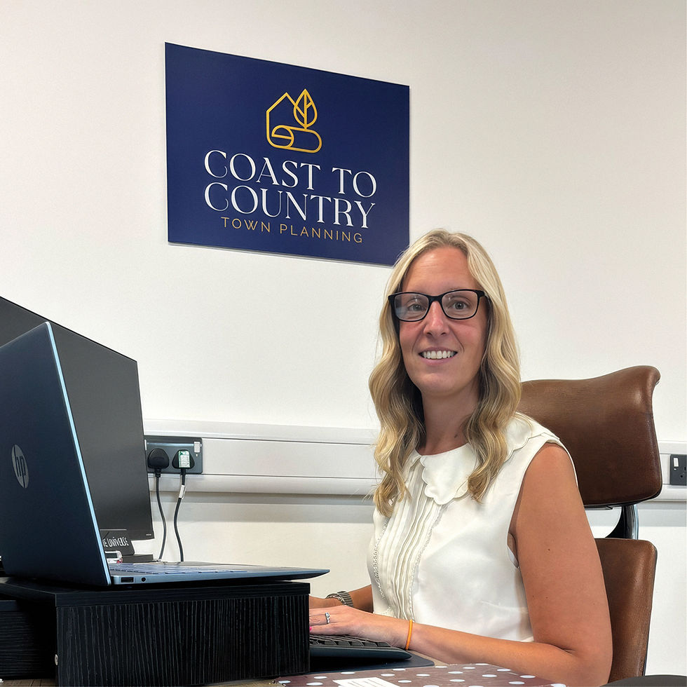 Rebecca Boyde Smiling woman with glasses sits at a desk using a laptop in an office; wall sign reads Coast to Country Town Planning.