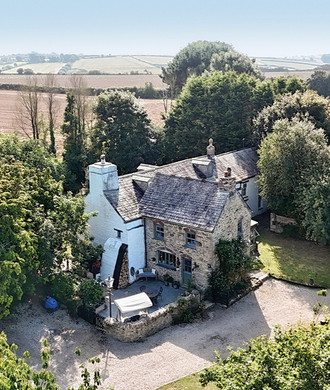 Aerial view of a stone farmhouse and outbuildings amid trees, gravel drive, and patchwork fields under a clear sky.