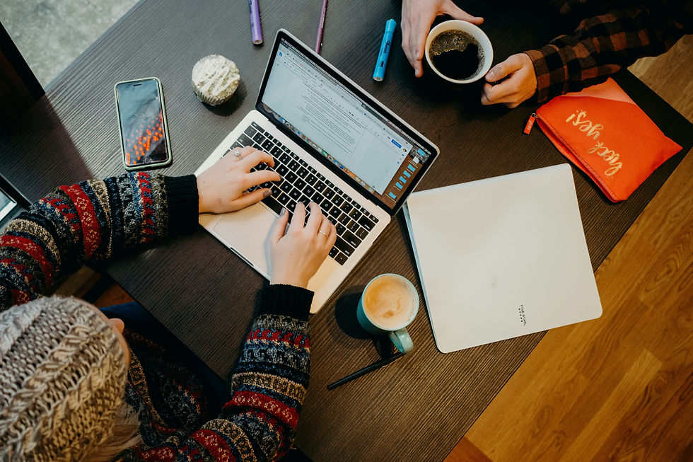 Overhead view of two people at a table; one uses a laptop, the other holds coffee. Colorful pens, a cupcake, and a phone on the table. Cozy mood.