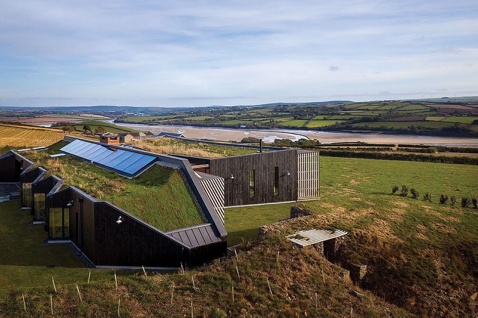 Modern eco-house with a grass-covered roof and solar panels, set in a green countryside overlooking a river and rolling hills under a blue sky.