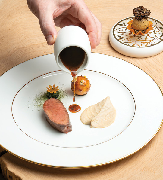 A hand pours sauce onto gourmet meat dish with herbs and gold-leaf potato on elegant white plate. Intricate dessert plate in the background.