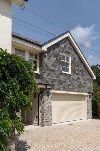 Modern house exterior with gray shingled gable, cream garage door, stone accents, blue sky, and leafy green shrubbery.