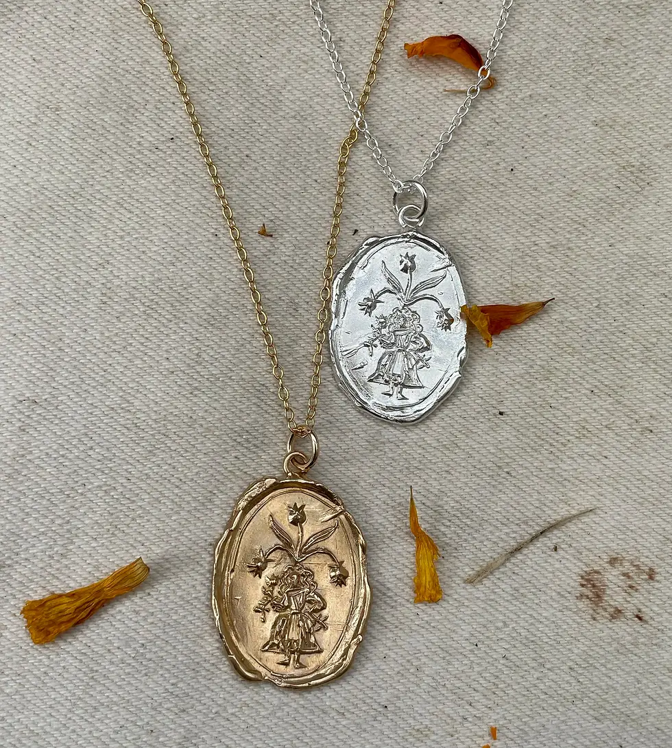 Two oval pendant necklaces, one gold and one silver, on textured fabric with scattered orange petals.