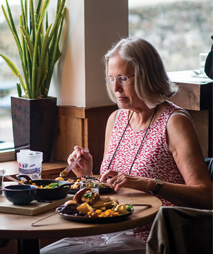 Woman in a red-patterned top eats at a wooden table with plates of food. A large plant and window are in the background. Calm mood.