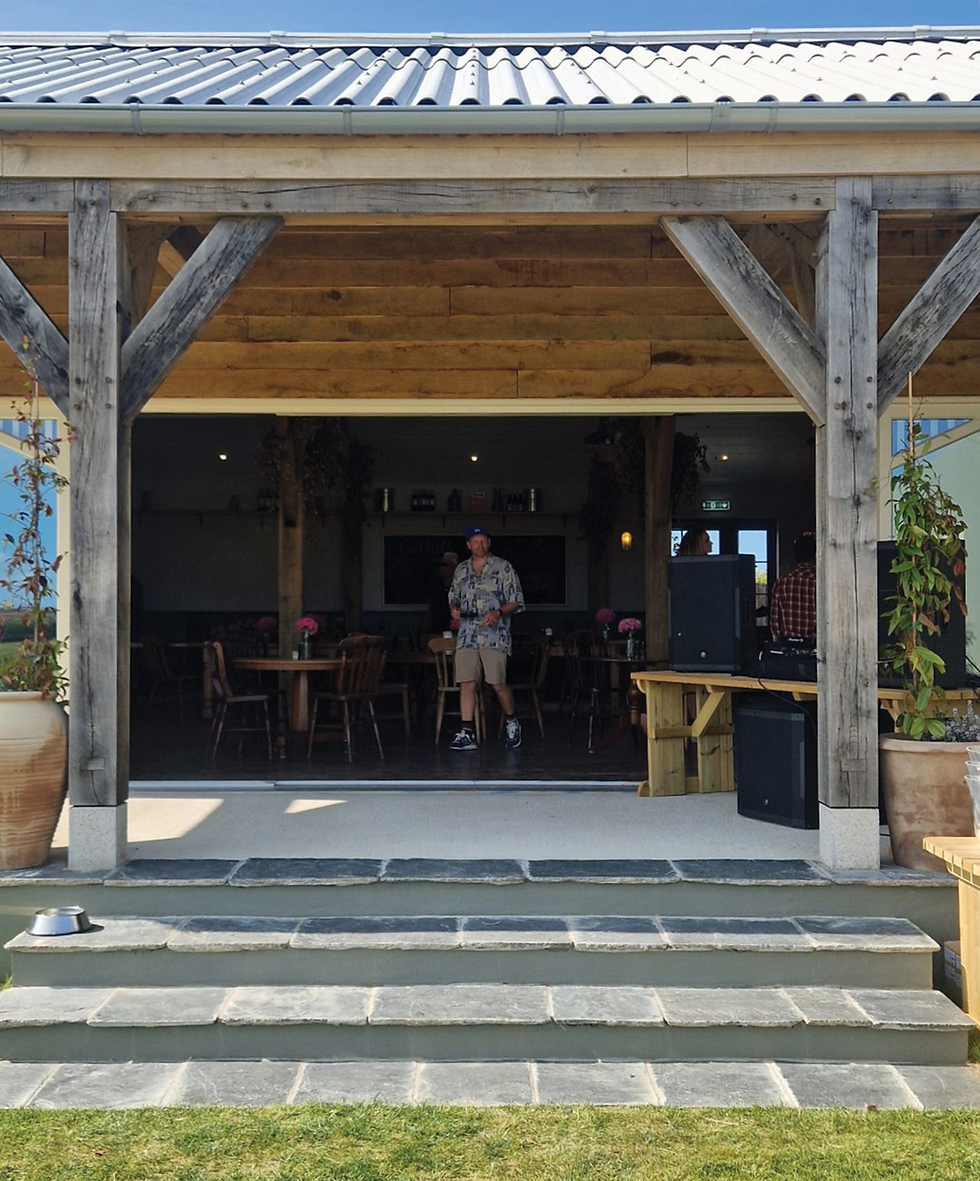 A person in a floral shirt stands in an open wooden pavilion with tables and pink flowers. Stone steps lead to a grassy area. Sunny day. the Halwyn entrance in the Post & Beam designed Oak frame building