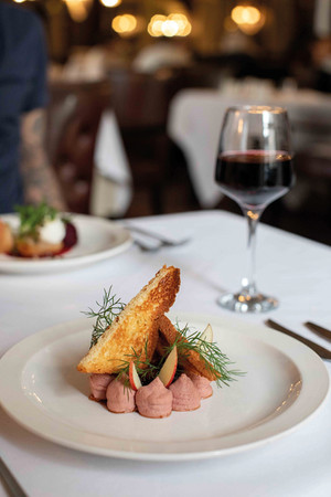Plate of pink mousse with toast slices, herbs, and apple slices on a white tablecloth. Glass of red wine in a dimly lit restaurant.