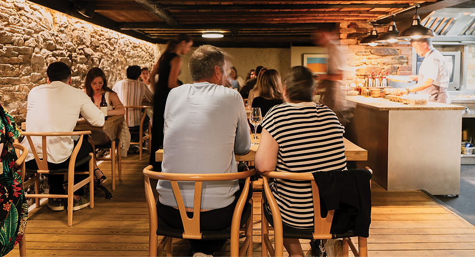 People dining in a rustic restaurant, seated at wooden tables. Stone walls and dim lighting create a cozy atmosphere. Chef works in background.