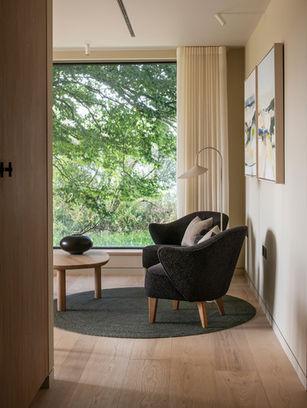 Modern reading nook with two dark armchairs, round table, and floor lamp beside a large window overlooking green trees.