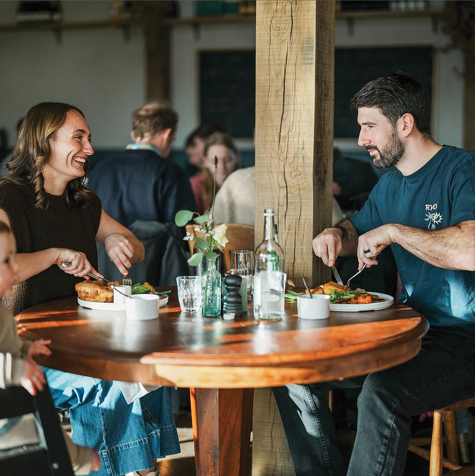 Couple dining at a wooden table in a restaurant, laughing and eating. Sunlight illuminates the scene. Other patrons are in the background.