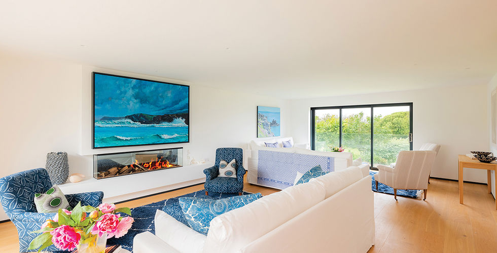 Bright modern living room with white sofas, blue chairs, ocean artwork above a fireplace, flowers, and garden view through sliding doors.