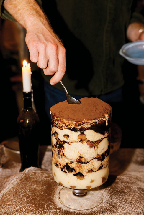 Hand serving layered tiramisu with a spoon, beside a lit candle on a table. Warm, cozy ambiance with dark background.