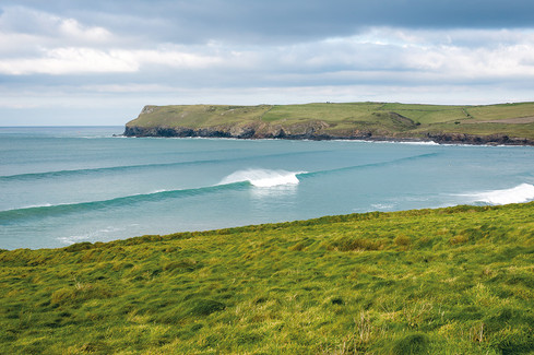 The view from St Moritz Hotel in Cornwall. Rolling waves approach a grassy coastline under a cloudy sky. Rugged cliffs line the horizon, creating a serene, natural vista.