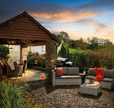 Cozy outdoor patio with wicker chairs under a wooden pergola, gray sofas, a grill, and a table set for dining. Sunset sky overhead.