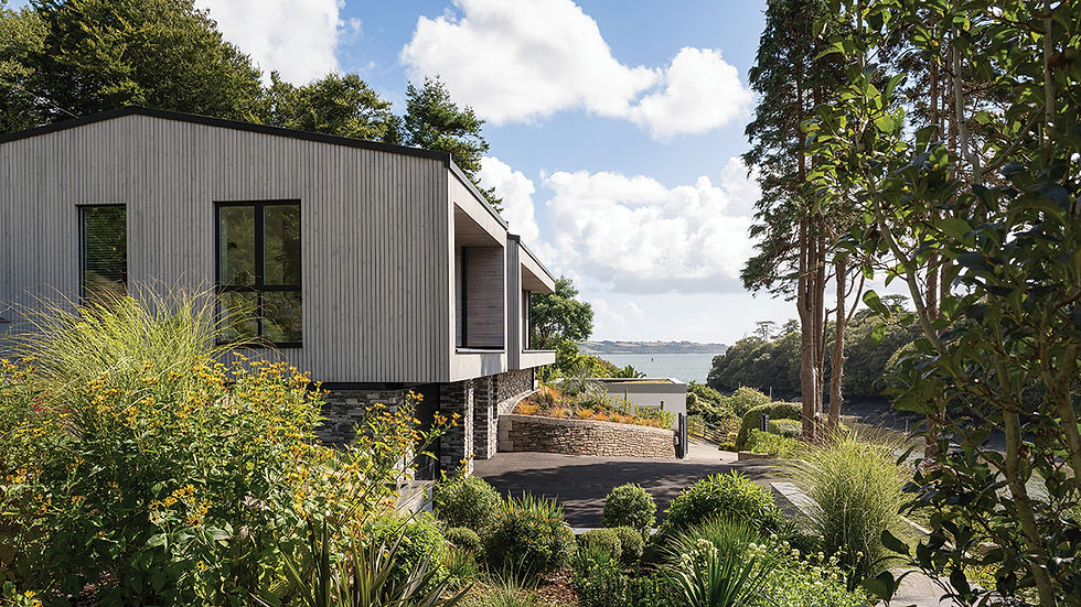 Modern house with lush garden overlooking the sea under a bright, cloudy sky.