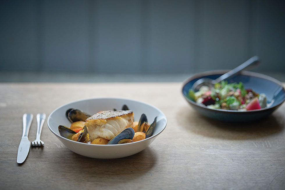 Plate of fish with mussels and potatoes on wooden table. Salad bowl with greens and radishes in background. Fork and knife beside plate.