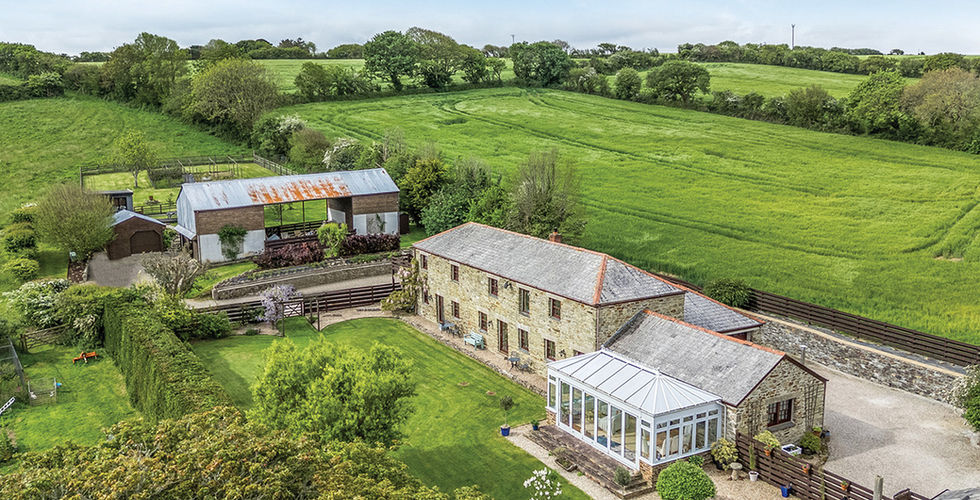 Aerial view of a stone farmhouse, barn and greenhouse beside bright green fields under a blue sky