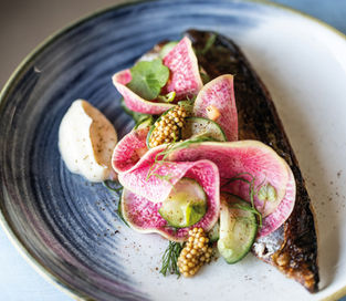 Grilled fish on a blue-rimmed plate, topped with radish slices, mustard seeds, greens, and a dollop of creamy sauce. Elegant and vibrant.