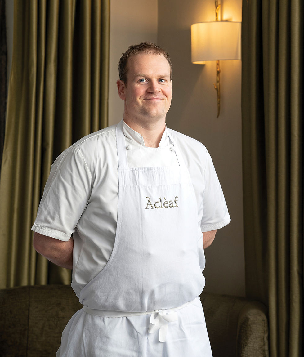 Chef in a white apron with "Àcleaf" text stands in front of green curtains, smiling in a warmly lit room. Calm and professional mood.