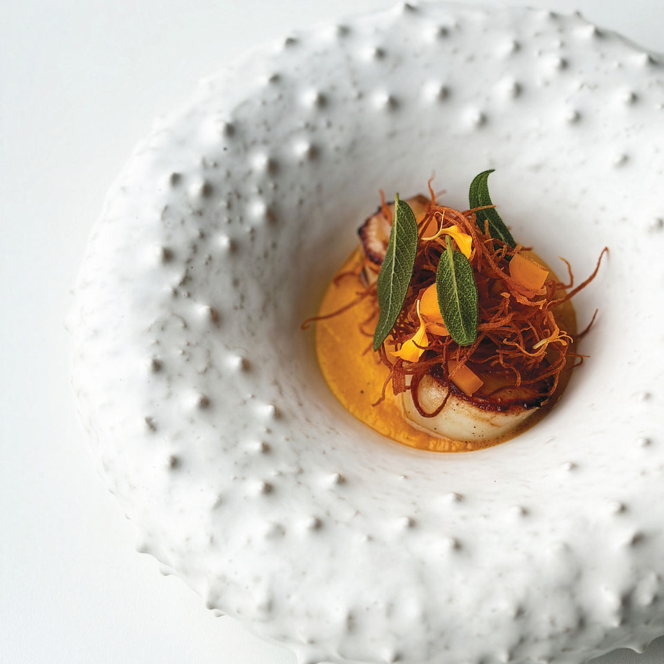 Gourmet dish in a textured white bowl, featuring seared scallop, orange sauce, and green herbs, creating an elegant culinary presentation.