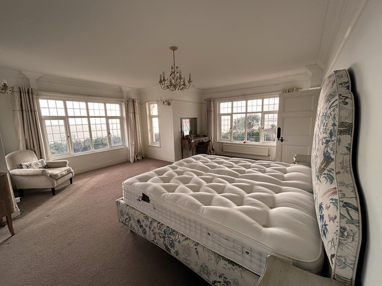Spacious beige bedroom with floral upholstered bed, armchair, vanity, and large windows under a chandelier, softly lit and tidy.